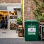 Contenitore verde sicuro per la raccolta dell’olio alimentare esausto nel cortile esterno di un ristorante moderno durante una giornata estiva.