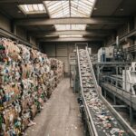Balle di rifiuti in plastica stoccate all’interno di un impianto industriale di riciclo con macchinari di selezione sullo sfondo